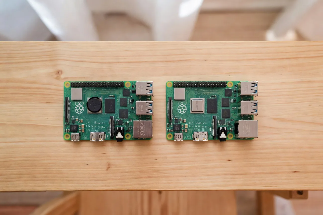 Raspberry Pi 5 and Raspberry Pi 4 boards side by side on a clean wooden desk, top-down view showing GPIO and ports